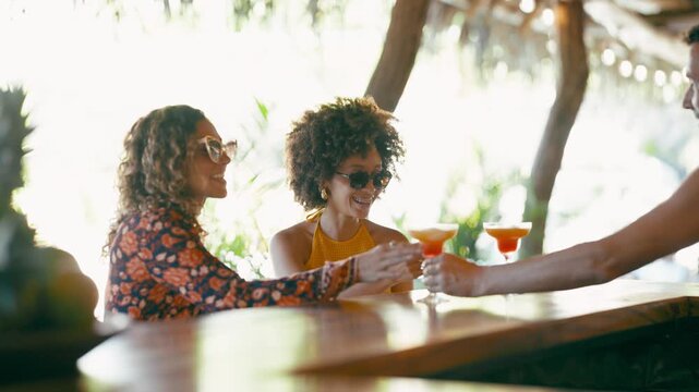 Two beautiful diverse women smiling and toasting with cocktails. A bartender serves colorful drinks to the happy friends at an outdoor beach bar