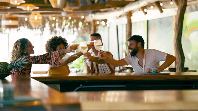 Group of four happy, diverse friends sitting at a wooden bar counter in a tropical themed bar, toasting and celebrating together