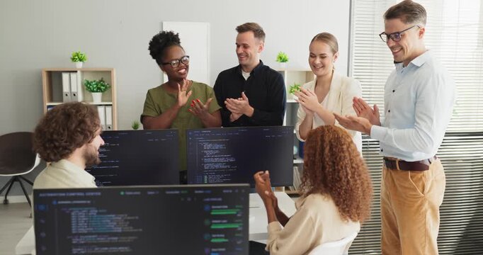 Programmers team developers in office share applause, celebration. They are clapping by monitors with coding visible, noting success and teamwork. Clear team celebration concept now.