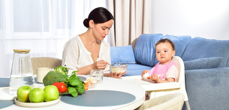 Mother feeding baby with a spoon. Spoon-feed. Happy young mother with a baby in the kitchen interior. Fresh vegetables and fruits.