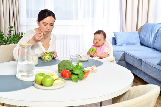 Mother feeding baby with a spoon. Spoon-feed. Happy young mother with a baby in the kitchen interior. Fresh vegetables and fruits.
