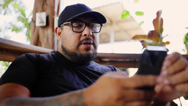 Latin man with a beard, glasses, and a hat scrolling and texting on his phone. He is sitting on a wooden bench at a steakhouse in Costa Rica