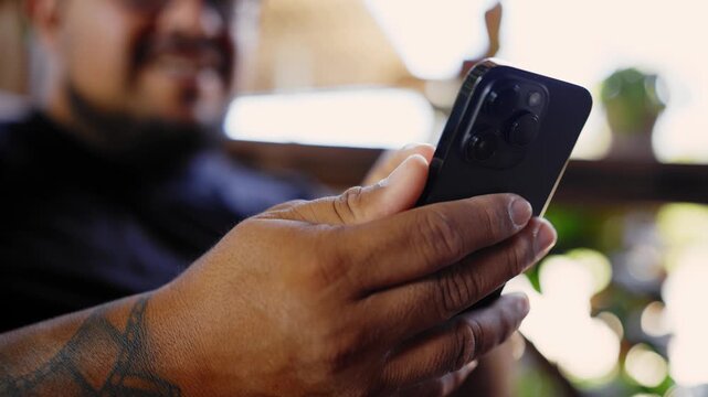 Close up of a man with tattoos on his arm, happily scrolling and typing on his mobile phone while sitting in a steakhouse restaurant