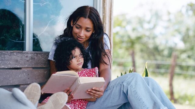 Loving Latina mother teaching her daughter how to read a book. They are sitting on a wooden bench outdoors, enjoying a quiet moment together