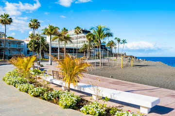 White benches and vibrant flower beds line a coastal promenade overlooking a volcanic beach sports area in Puerto Naos town, La Palma, Canary Islands, Spain © pkazmierczak