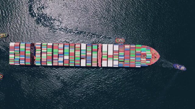 Aerial top-down view of a massive container ship loaded with colorful cargo containers being maneuvered by several powerful tugboats on the open ocean for international trade and shipping
