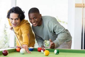 Obraz premium Diverse male friends in mustard sweater, light sweatshirt playing on green-felt table in game room
