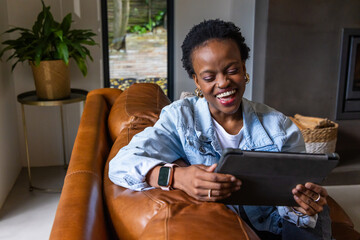Obraz premium African American woman sitting on sofa in living room wearing denim jacket holding tablet smiling