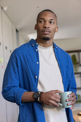 African American man standing in kitchen near cabinets holding pale green mug, wearing smartwatch © wavebreak3