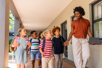 Fototapeta premium Diverse children wearing backpacks walking along school corridor with female teacher holding tablet