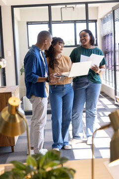 Diverse coworkers discussing plans at office corridor wearing smart casual with laptop, papers