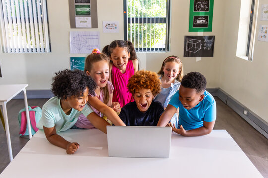 Diverse children leaning around open laptop in classroom, pointing at laptop screen and backpack