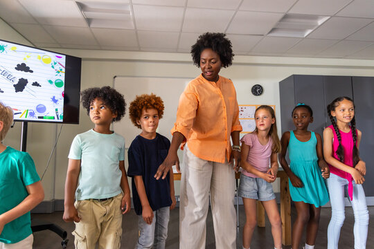 African American teacher in orange blouse guiding children at front of class with monitor