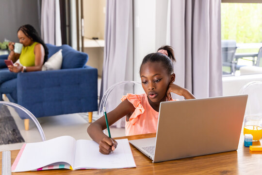 African mother holding phone and mug while youth girl writing in notebook at table with laptop