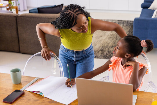 African mother leaning over youth daughter guiding homework at table with laptop and lime top