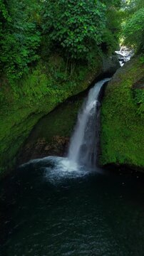 Aerial view of a hidden waterfall deep within the lush tropical rainforest of Mount Daun, Indonesia. Surrounded by dense green jungle vegetation, the pristine cascade flows into a dark natural pool