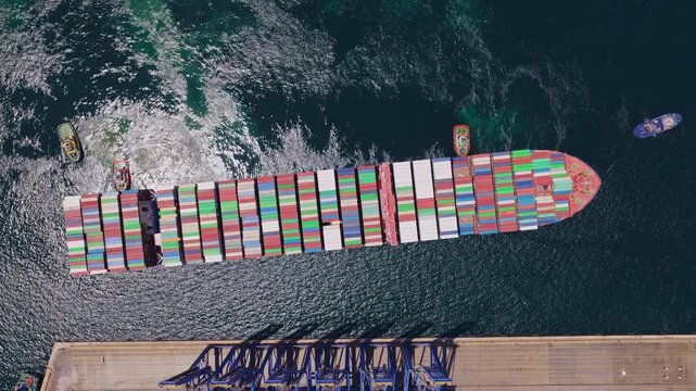 Impressive top-down aerial footage capturing a massive cargo vessel loaded with colorful containers being expertly maneuvered into a commercial harbor by a fleet of assisting tugboats