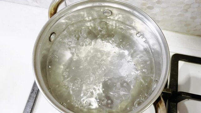 Water intensely boiling in a stainless steel pot on a gas stove burner, bubbling and steaming as it heats up for cooking, top view close up of the simmering liquid