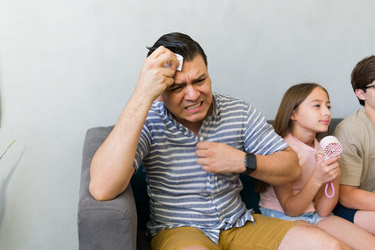 Man and family suffering from extreme summer heat indoors, father sweating and daughter using a personal fan