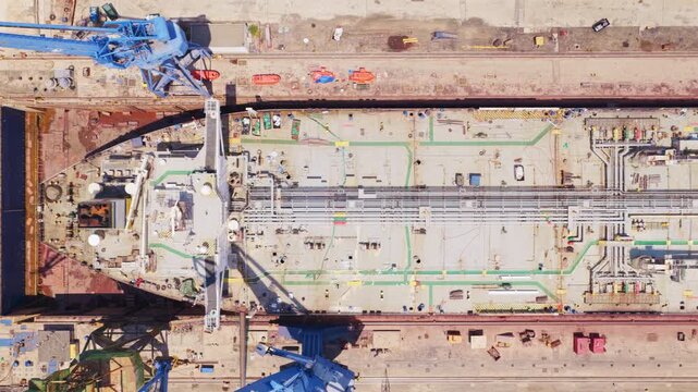 Aerial top-down view showing a massive tanker ship under construction or repair inside a huge dry dock facility, with large gantry cranes moving alongside the vessel during maintenance work