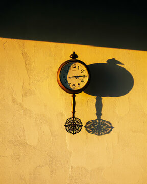 Round clock with Arabic numerals and decorative pendulum on a textured wall with sunlight and shadows