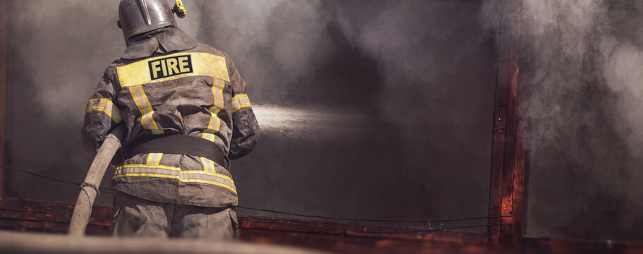 Firefighter spraying water on burning building, heavy smoke and flames, emergency firefighting action, back view of fireman in protective gear during dangerous fire rescue operation