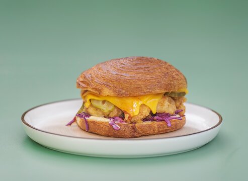 Golden croissant holds tender chicken fillet topped with rich cheddar cheese, crunchy pickles, and vibrant purple cabbage. Perfectly placed on a simple white plate, this meal invites indulgence