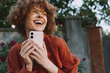 Joyful woman outdoors holds a phone, smiling with curly hair in a red sweater. Bright natural light...