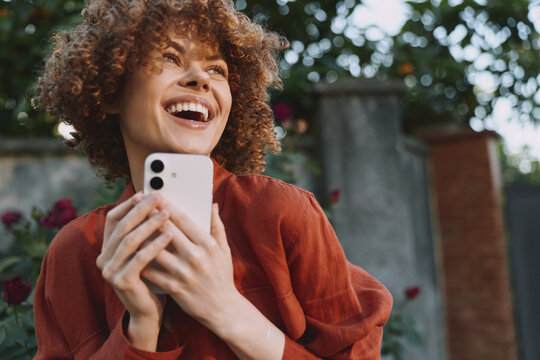 Joyful woman outdoors holds a phone, smiling with curly hair in a red sweater. Bright natural light and greenery frame a candid lifestyle moment focused on happiness and everyday joy