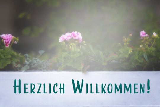 Herzlich Willkommen auf Schild in wei&szlig; mit Blumen im Hintergrund