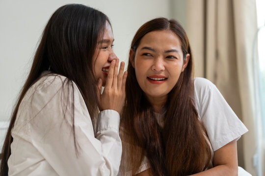 Two Asian women laughing and chatting together on bed in cozy bedroom, symbolizing friendship, communication, and positive lifestyle with smartphone interaction, casual clothing, and warm domestic