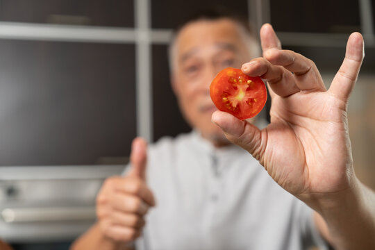 Elderly man covering eye with tomato in playful concept, representing healthy lifestyle, nutrition, and wellness in cozy indoor kitchen environment with natural daylight, vegetables, and cheerful