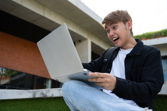 Young Asian graduate celebrating success with laptop outdoors in front of modern building, symbolizing achievement, startup growth, and positive career lifestyle with joyful expression, technology