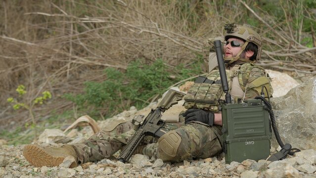 A special forces soldier rests and rehydrates in a rugged environment, awaiting orders on his tactical radio during a demanding military mission.