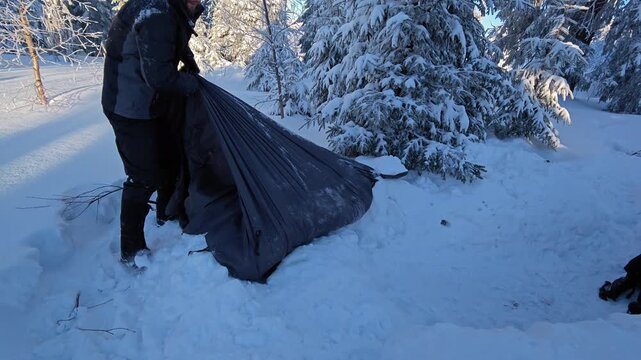 Survivalist pulling heavy black tarp shelter covered in snow in winter pine forest