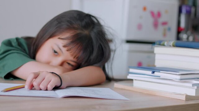 Close up selective focus of demotivated Asian kid girl resting her head on a desk in daylight. She looks overburdened by too much homework, illustrating academic pressure and childhood study burnout.