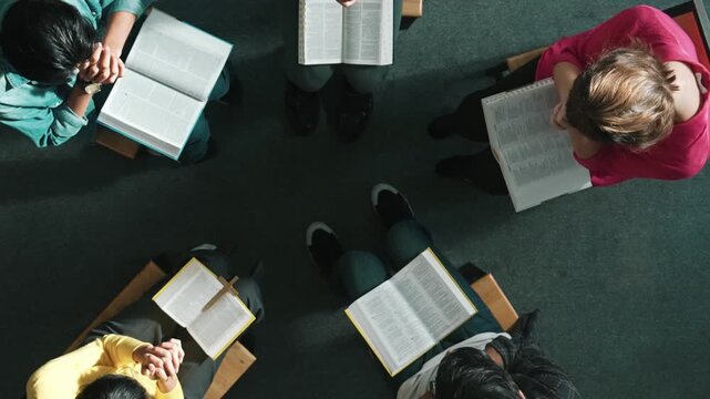 Top down view of prayers reading bible with cross on the laps while making folded hands. Aerial view of diverse people praying to god with faith, trust and hope. and sitting in circle. Symposium.