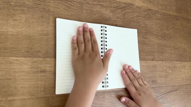 Child Hands Smoothing the Left Page of a Small Spiral Notebook from an Oblique Overhead View, 4K Silent Video