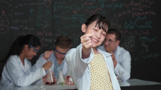 Asian scientist show miniheart to camera while diverse group doing experiment. Young schoolgirl standing at blackboard with chemical theory written while wearing lab coat at laboratory. Edification