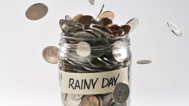 Close-up shot of coins falling in slow motion into a coin jar labeled 'Rainy Day'.
