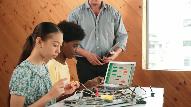 Teacher standing while explain coding and robotic construction. Diverse teenager fixing mother board and using electronic tool while looking at laptop display programming code screen. Edification