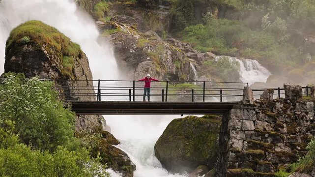 Woman hiker crosses a bridge over the Kleivafossen waterfall on the way to Brikdalsbreen Glacier in Norway.