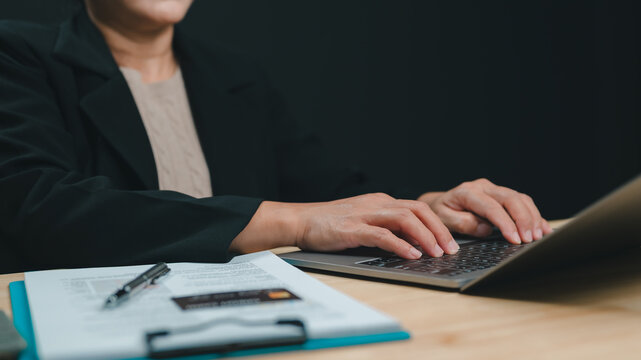 Professional businesswoman wearing black formal blazer types laptop keyboard beside clipboard pen credit card nearby against dark background indoors evening office work session performing financial.
