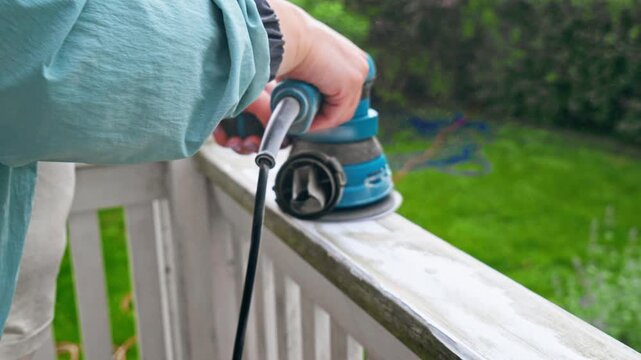Electric sander smoothing white painted wooden railing outdoors, close up of hands using power tool