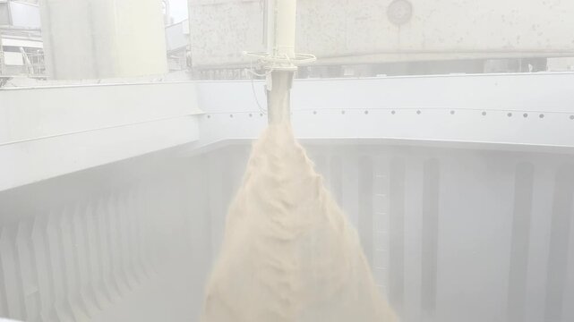 Grain terminal operation. wheat flows from chute into ship hold, creating massive dust cloud. Bulk loading of dry cargo for international shipping.