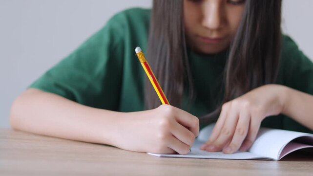 Close up selective focus of a concentrated Asian kid, a young Thai girl or Chinese student, diligently doing homework and writing with a pencil in a notebook. Education, learning, and study concept.