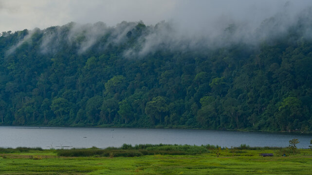 Scenic view of Lake Buyan in Bali with dramatic low clouds and mist rolling over a lush green tropical rainforest mountain landscape.