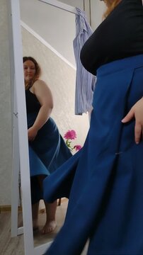 Plus size woman spins in front of a mirror, checking her long flowing blue wrap skirt at home. Low angle vertical mirror shot. Body positivity and dynamic fashion lifestyle concept video.