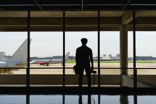 Silhouette of a male photographer holding a camera, standing by a large window at an airport terminal and looking out at airplanes.