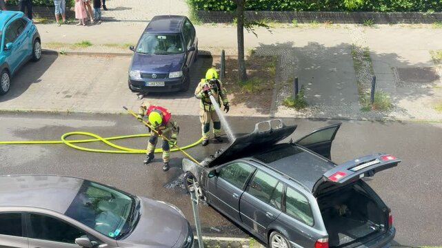 Two firefighters control a burned station wagon on a city street. The open hood, trunk door, hoses and smoke mark a dramatic vehicle fire emergency.